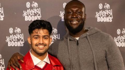 Sufiyaan Salam pictured with Stormzy after he won the New Writers' Prize. Sufiyaan is a 26-year-old British Asian man with his dark hair long on top but shaved close on the sides. He has brown eyes and a short beard and moustache. He wears a red leather jacket over a red jumper and white shirt. Stormzy stands about a foot taller to his left. The rapper is a black man in his 30s with cropped hair and a short beard. He has his arm around Sufiyaan's shoulders and wears a grey hoodie. They're photographed in front of #Merky Books event hoarding