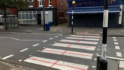A pedestrian crossing which is white has had red crosses painted in each of the sections. Behind the crossing is a closed restaurant.