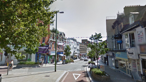 A road junction with a number of shops either side of a road with trees either side of the road.