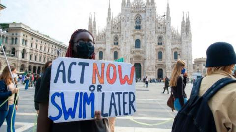 A climate protester holds a sign reading "Act now or swim later" in Milan