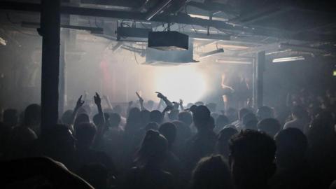 Silhouetted figures are shown dancing with arms aloft in front of a bright white light inbetween pillars in Sankeys Soap in Manchester