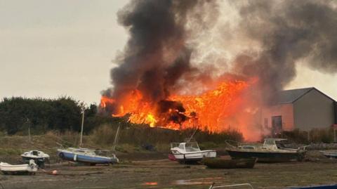 Large orange flames and thick smoke rise into the sky from the structure which is on fire. The photo has been shot from the opposite bank of a river at low tide. Several small boats are in he foreground.
