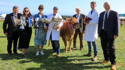 A group of seven men and women holding trophies and smiling in a field along with a brown cow which has a rosette attached to its harness.