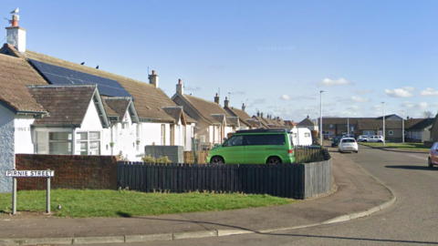 A residential street with white houses, grey rooves and cars parked out front. A street sign reads Pirnie Street