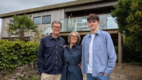 An older couple, a man and a woman, stood outside their home with their son. The couple are wearing navy outfits, and the young lad is in a light blue shirt, top and jeans. The wooden house is on stilts, with a large glass balcony seen on the first floor.