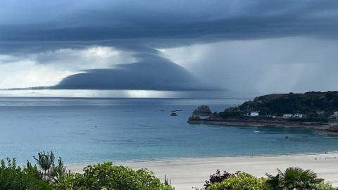 The sky has a top layer of dark blue cloud with a 'hat' shaped large cloud below, then sea, a headland to the right and beach in the foreground