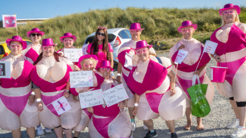 A cheerful group of people is gathered outdoors on a sunny early evening.  They are all dressed in inflatable pink and beige costumes with bright pink hats, sunglasses, and various hand held signs promoting breast cancer awareness. Some of the signs read “No one fights alone,” and “Big and small, save them all.” 
