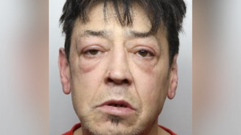 A police mugshot of a dishevelled looking man. He has eye bags and puffy eyelids. He has a bit of hair below his mouth but is mostly clean shaven. He has messy black hair around his forehead.