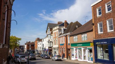 Sevenoaks High Street in Kent, England. Several stores are visible such as Barclays Bank in the centre and Savills Real Estate Office on the right. There are also people in the background.