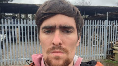 Luke Jones has brown hair and eyebrows and is standing in front of an iron gate.