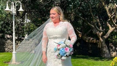 Stead in white wedding dress and veil on lawn holding bouquet