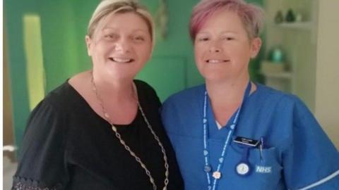 A woman in a black top stands next to a woman in blue scrubs and a name tag in a birthing room.