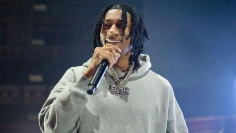Digga D on stage wearing a grey hoodie and a necklace holding a black microphone to his mouth. He has long, braided black hair.