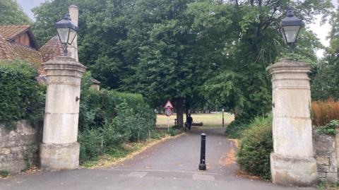 Two stone gateposts standing either side of a walkway entrance into a park. The gateposts have old-fashioned street lights on top and a bollard and sign can be seen with the words "10 max" written on it. Trees and bushes can also be seen.