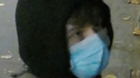 A CCTV image showing the person, who has curly brown hair, wearing a black hoodie and blue facemask