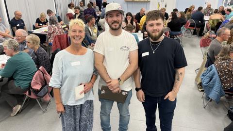 Organisers Kate Linnegar, Jamie-Lee Crabtree and Ed Colton stood together in front of a people sat around tables at the Broadgreen Community Centre in Swindon