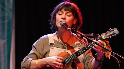 A woman with dark hair with her eyes closed sings into a microphone while playing a ukulele