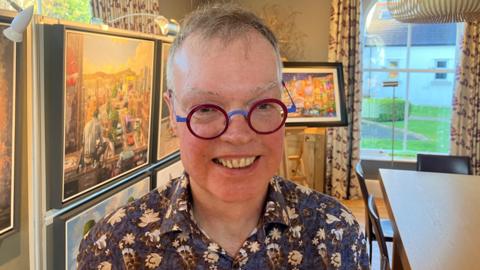 A man smiling at the camera. He is wearing a blue and white patterned shirt and red and blue glasses. Behind him are some paintings under display lights.