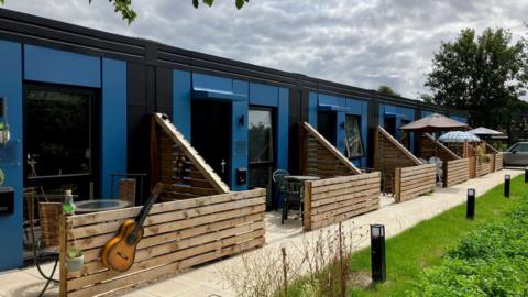 Front of a blue coloured building. There are seats and tables outside of the doors with a wooden panel in front of them. There is also a guitar stuck to the front of the wooden panel closest to the camera.