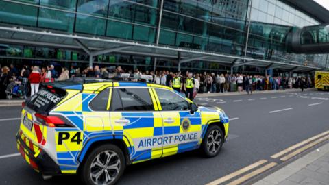 A busy picture, with an airport police car in the foreground. There are two people in high-vis. The back of another emergency vehicle can be seen at the top right. There is also a blurry queue of people lined up outside the glass terminal building.