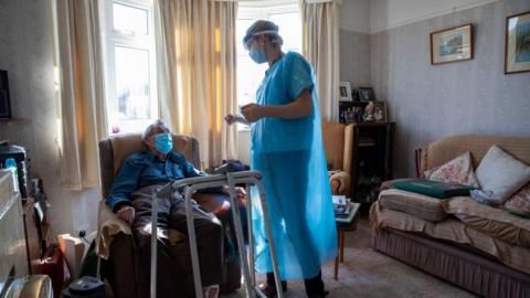 A healthcare worker standing and dressed in a blue gown chats to a housebound patient in his front room. The patient is sat down on a chair with a medical walking frame in front of him. He is wearing a mask. There is a sofa on the right of the picture and sun is coming through the windows behind.