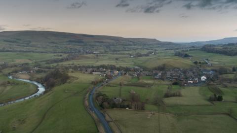 This is an overhead shot of Bainbridge, in the Yorkshire Dales. A river is winding through the left of the shot, the village is on the right, the hills are in the background, a dark and threatening sky above. North Yorkshire Council says the size - and rural nature - of the area it serves adds extra costs to its services.