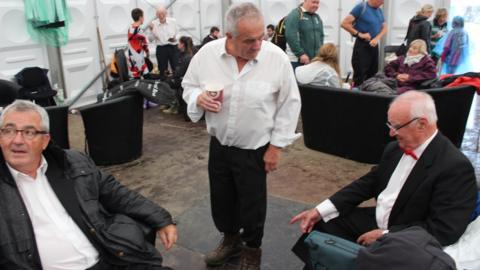 Aelod o gor yn pwyntio tuag at esgidiau mwdlyd ei gyd-aelod // A choir member points towards his fellow choir member's muddy boots