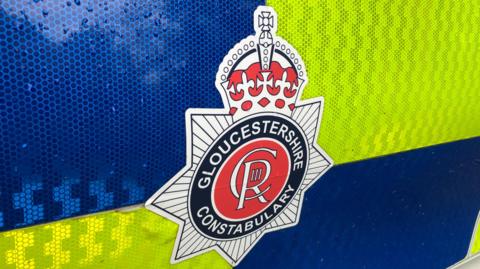 A Gloucestershire Constabulary badge on the side of a yellow and blue police car door. It is a red circle with a black outline and a crown on top of it. Gloucestershire Constabulary is written around the circle in white text.