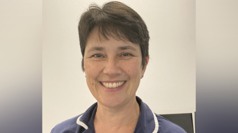 Lucie Legg wearing a blue nurse's uniform with white trim. She has short brown hair and is smiling.