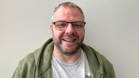 A man in glasses, with a short beard and moustache smiles at the camera. He is wearing a green hoodie over a grey T-shirt