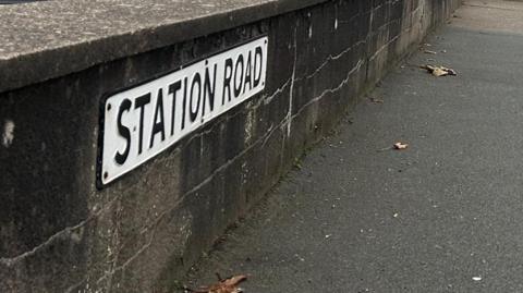 A black-and-white road sign for Station Road attached to a low brown wall with a tarmac pavement running alongside it.