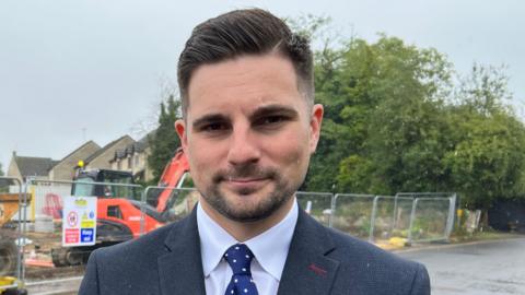 Joe Harris, a man with neat brown hair and a brown beard, stands on the pavement beside a wet road, with a construction site and houses behind him.