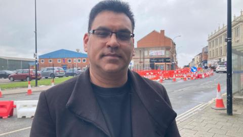 An Asian man in a smart dark jacket and glasses stands in front of a road with roadworks