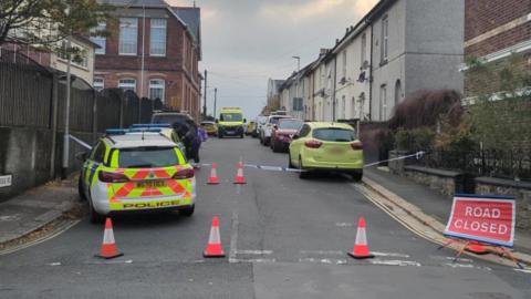 Two police cars and a police officer at a taped off road with a red road closed sign while an investigation takes place. An ambulance and several cars are parked on the road.