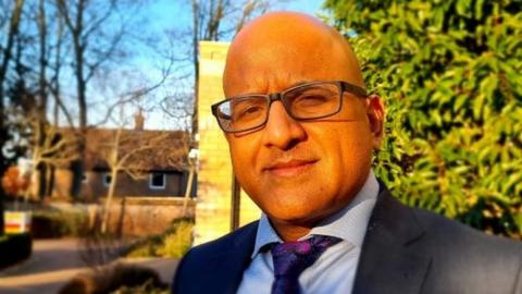 Sanjay Kaushal dressed in a suit, wearing glasses, standing outside in daylight. He owns six care homes and is a director of the Norfolk Care Association