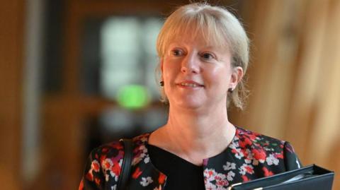 Shona Robison, with blonde hair and wearing a black top and patterned jacket, walks in the Scottish Parliament, carrying a black folder.