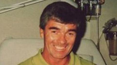 An old picture of a man in a green shirt and sat in a white armchair while smiling in the direction of the camera