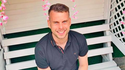 Gavin Campbell is wearing a navy blue polo top while sat on a white bench. He has light stubble and short brown hair.