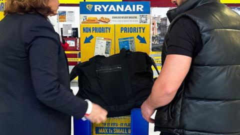 Two unidentified passengers, a man and a woman wearing black, attempt to put a large black bag into a Ryanair bag measurement box. It doesn't look like it fits. The box is in Ryanair's blue and yellow colours, with the words "priority" and "non-priority" with arrows. 