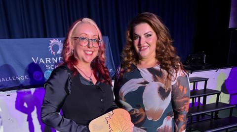 Jolie Bartley, Head of Performing Arts at Varndean School, pictured with West End star Jodie Prenger. The pair are pictured at Varndean School and Jolie is holding her award. Jolie is wearing a black top and has blonde/red dyed hair. Jodie is wearing a flowered dress and has long brown hair.