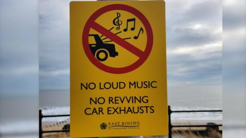 A yellow street sign with an illustration of a car. The text reads no loud music and no revving car exhausts.
