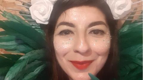 Sheetal smiling into the camera with dark hair and eyes and red lipstick, glitter on her face, white roses around her head and green feathers behind her head