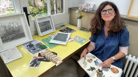 Sophie Molins sitting at a desk surrounded by framed artworks, photos and newspaper clippings and holding a book containing cut-out images