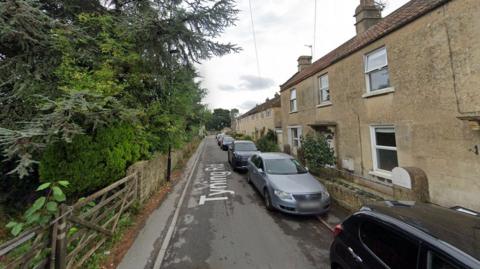 Google maps image of Tyning Road in Combe Down, Somerset. It is a narrow street with houses down one side and cars parked along the road. The other side of the street has a wall and lots of trees. 