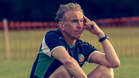 Steve Coogan sitting on the sidelines in a Republic of Ireland training kit looking disconsolate. He has grey hair and has his left elbow leaning on his knee.