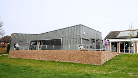 A grey, single storey building on an elevated brick platform. Some baloons can e seen by the side entrance of the property.