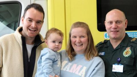 Tim, Baby Cecilia, Alex and EEAST paramedic Mike Sage. A man wearing a cream coat is standing on the left next to his wife Alex who is wearing a blue jumper and holding her daughter. Mike who is a paramedic is standing next to them. The group pf people are standing in front of a Ambulance