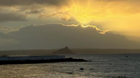 A picture of a large body of water, with an island in the distance. Above the water the sun can be seen breaking through the clouds, causing a yellow glow.
