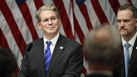 US Treasury Secretary Scott Bessent smiles sitting in front of a microphone and a backdrop of American flags while US Trade Representative Jamieson Greer is in the background