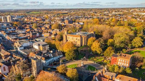 An aerial shot of Colchester with the castle in the centre of the picture. There is trees of a park surroudning it and housing and some shops in the distance. The sun light is slowly setting on the city.